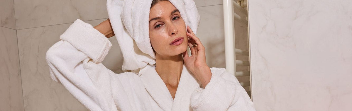 Woman with a towel wrap in a bathroom beside the glowing LumaLux Face V2 LED red light mask